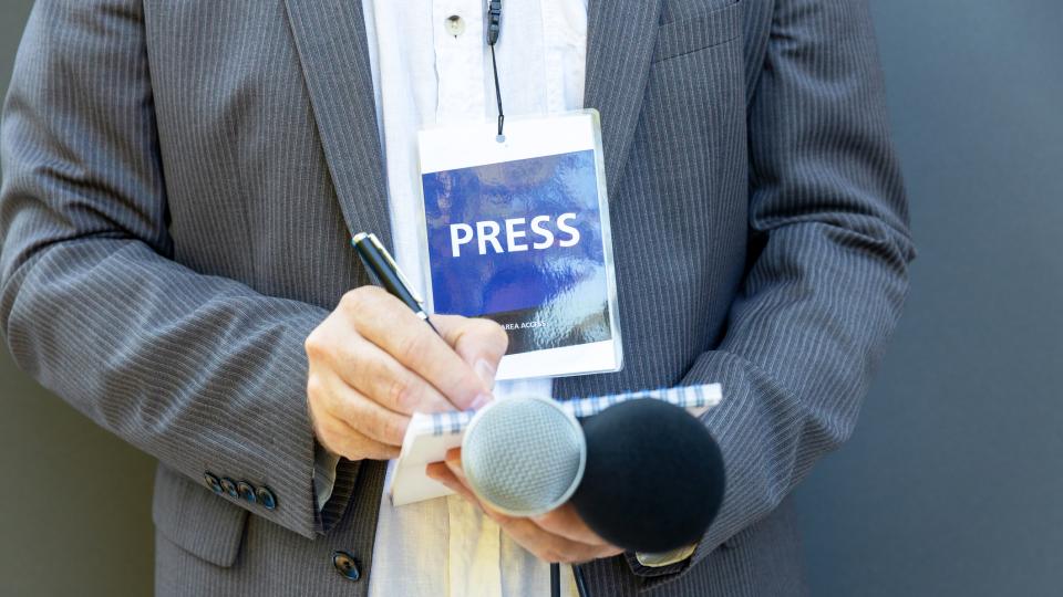A journalist wearing a press pass, holding a notepad and pen